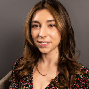 A close up headshot shows a young brunette woman in professional attire, smiling at the camera, set against a grey background that suggests an indoor setting.