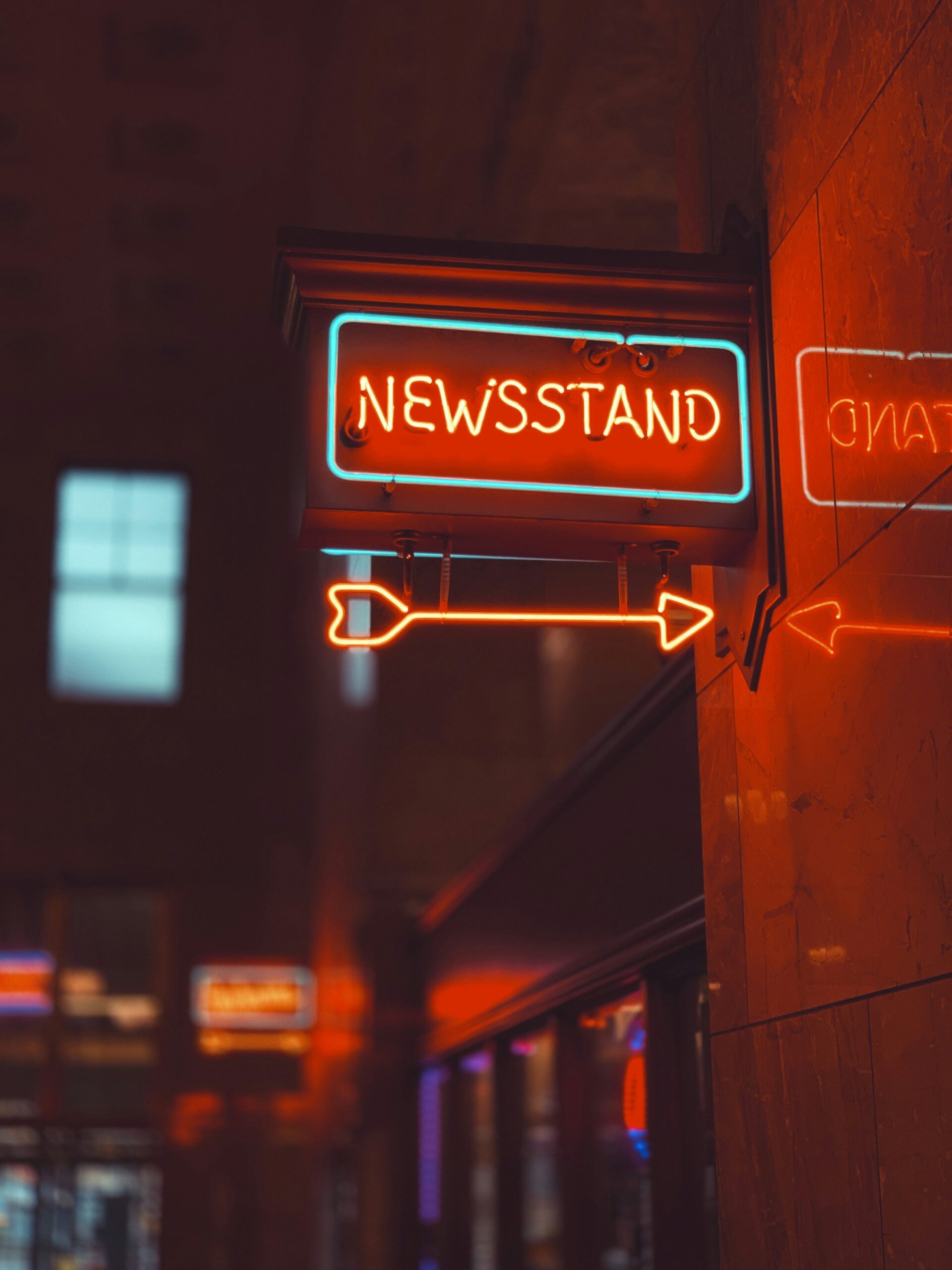 Stock photo showing a neon sign reading "Newsstand" in red, with a red neon arrow underneath. Darkened background and street signs out of focus suggest an outdoor nighttime setting.
