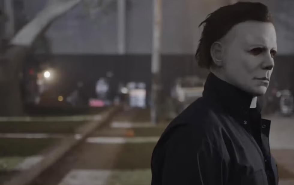 Image of a man with a white mask over his face (Michael Myers from the Halloween movie franchise), appearing to stare at something to his left