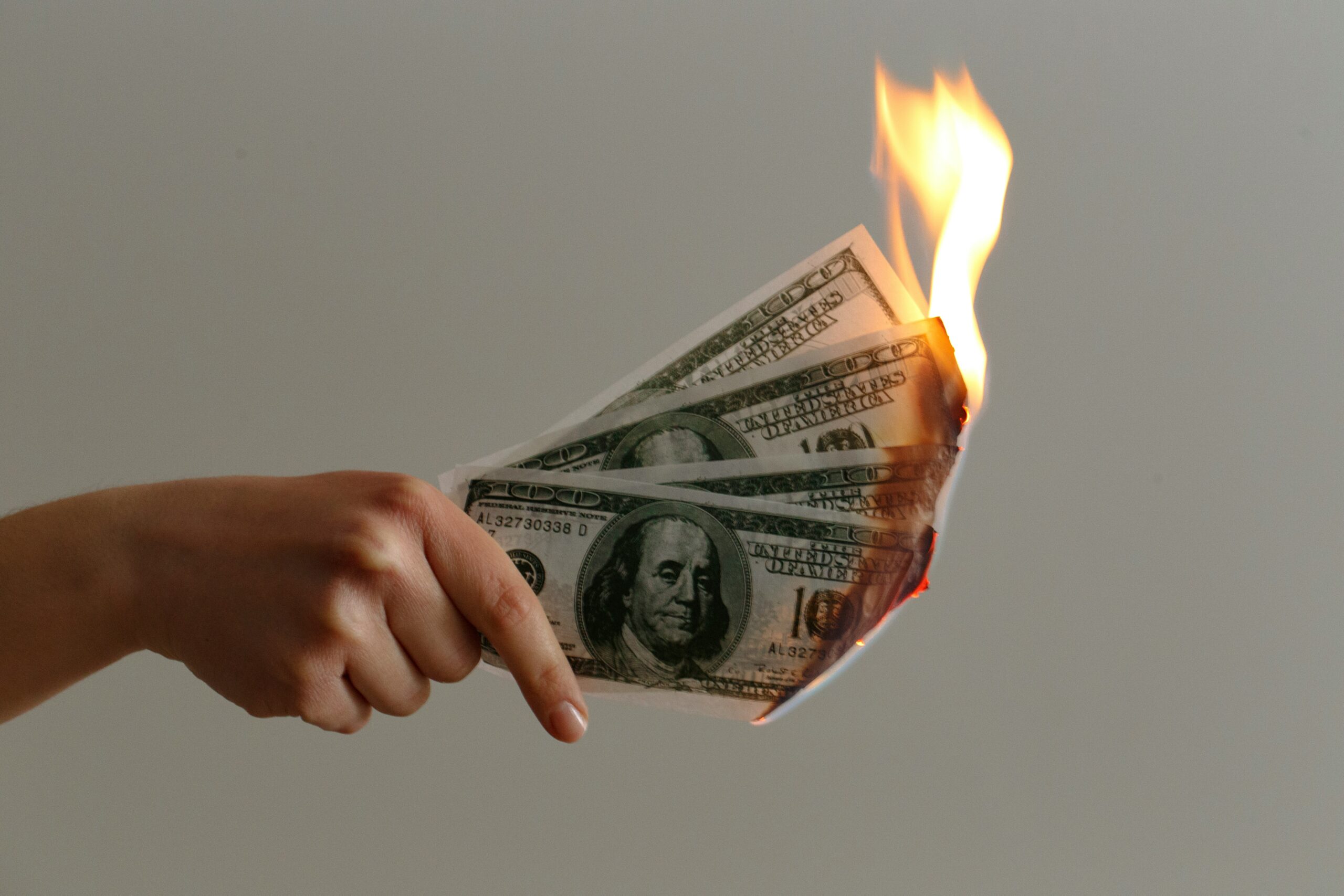Stock image of a hand holding several hundred dollar bills, which are on fire